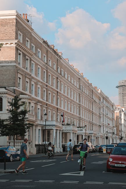A wide view of a row of classic four-storey Victorian terraced houses with ornate white facades, large sash windows, and balconies, situated along a busy street in South Kensington under a partly cloudy sky. On the pavement, a man with a backpack and a cyclist are seen riding past, while pedestrians are walking along the sidewalk. Several parked cars are visible adjacent to the buildings, and in the background, a construction crane indicates ongoing development. This scene captures the typical architecture and lively atmosphere of a central London residential area, aligning with house removals and furniture transport services offered by Man with Van South Kensington, who manages home relocation and packing processes in this affluent neighbourhood.