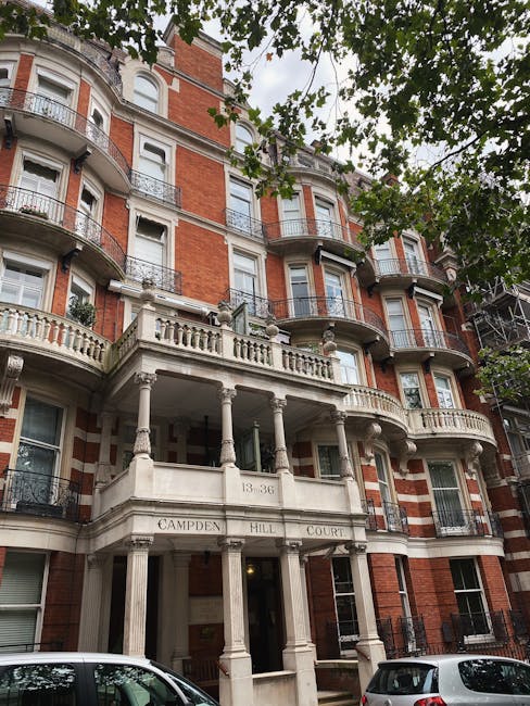 A multi-storey residential building with a red-brick façade and white decorative stonework, including rounded bay windows and wrought iron balcony railings, situated on a tree-lined street in South Kensington. The building's main entrance features a portico with tall, white columns supporting a curved balustrade, and the address number 13-36 is displayed above the doorway. In the foreground, two parked cars are visible on the pavement, and the scene is shaded by foliage from overhanging trees, with the sky partly visible through the leaves. The image captures the exterior of this historic home, which might be part of a house removal or relocation process, with some white plastic wrapping around furniture or boxes possibly visible nearby, indicating packing activities by a professional removals team such as Man with Van South Kensington involved in furniture transport and home relocation services in the area.