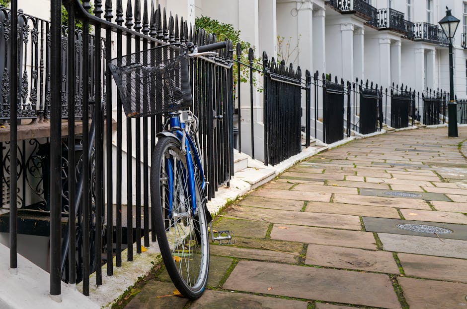 A cobbled street in South Kensington lined with residential buildings, some with potted plants and greenery along the pavement. In the foreground, the street appears empty, but further down, two people are visible walking away from the camera. The buildings on either side are a mix of modern and traditional architectural styles, with trees and bare branches overhead indicating a season outside of summer. The scene includes a loading bay area adjacent to a building with a blue exterior and metal railing, referencing Brompton Road loading bay rules for local removals. This outdoor setting captures an environment suitable for home relocation activities, with space for loading and unloading furniture and boxes as part of a furniture transport or packing and moving process, consistent with professional removals services by Man with Van South Kensington.