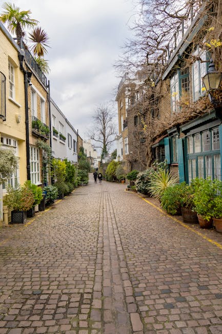 A narrow cobbled residential street on Gloucester Road with multi-storey houses on both sides, featuring a mix of brick and painted facades, some with large windows and balconies. The street is lined with potted plants and small shrubs along the pavement edges, creating an inviting atmosphere. In the distance, two people are walking away from the camera, while the overcast sky above casts a soft, diffuse light. The scene is typical of South Kensington, with the street's confined width and historic character. This setting presents typical challenges for house removals on tight access streets, where careful planning and the use of appropriate equipment are necessary. Man with Van South Kensington specializes in home relocation and furniture transport within such narrow environments, ensuring safe and efficient delivery of belongings from the property to a moving vehicle, possibly visible at the end of the street, where loading processes are underway.