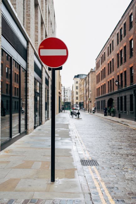 A red and white 'no entry' traffic sign mounted on a black pole positioned on a paved sidewalk adjacent to a narrow urban street. The sidewalk, constructed of light-colored stone slabs, runs alongside multi-story brick and stone buildings with large windows. In the background, there are a few pedestrians and cyclists, with some carrying bags or riding bikes, as well as parked bicycles leaning against railings. The street appears slightly wet, possibly after rain, and features a double yellow line along the curb, indicating no parking. The scene is illuminated by natural daylight with an overcast sky, and the overall setting suggests an inner-city area suitable for home relocation and furniture transport movements. Occasionally, Man with Van South Kensington’s services may be involved in assisting moves through tight street access areas in South Kensington, such as Gloucester Road.