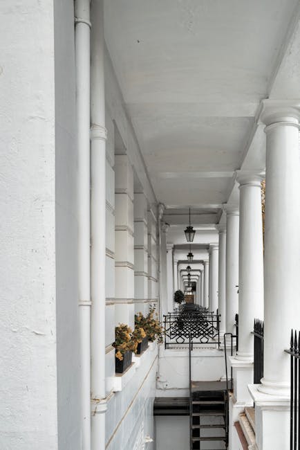 A narrow cobbled residential street on Gloucester Road with multi-storey houses on both sides, featuring a mix of brick and painted facades, some with large windows and balconies. The street is lined with potted plants and small shrubs along the pavement edges, creating an inviting atmosphere. In the distance, two people are walking away from the camera, while the overcast sky above casts a soft, diffuse light. The scene is typical of South Kensington, with the street's confined width and historic character. This setting presents typical challenges for house removals on tight access streets, where careful planning and the use of appropriate equipment are necessary. Man with Van South Kensington specializes in home relocation and furniture transport within such narrow environments, ensuring safe and efficient delivery of belongings from the property to a moving vehicle, possibly visible at the end of the street, where loading processes are underway.
