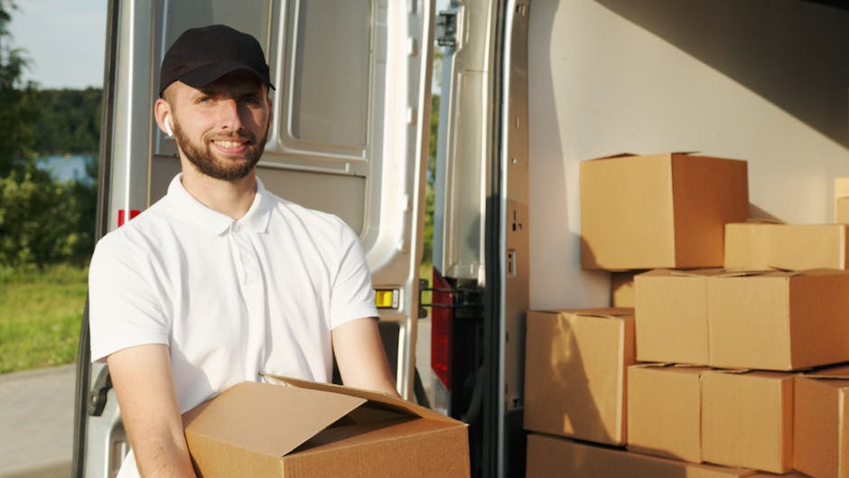 A man with a beard, wearing a black cap and a white polo shirt, stands outdoors next to a white moving van during daylight. He is holding a cardboard box in front of him, prepared for transportation as part of a house removal or furniture transport process. The van's rear doors are open, revealing a stack of similar cardboard boxes inside, along with plastic wrap and packing materials. The van is parked on a paved area beside grass and trees, with clear weather conditions and natural light illuminating the scene. This loading process is part of a professional home relocation effort, supported by Man with Van South Kensington, involving packing, loading boxes, and transporting household items.