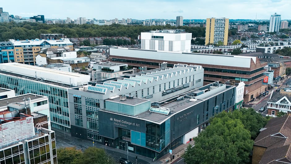 An aerial view of the Royal College of Art building in South Kensington, showing modern glass and concrete architecture with flat roofs, some featuring skylights and HVAC units. Surrounding the college are neighboring commercial and residential buildings, with a few trees visible at street level. The scene is taken during daytime with clear skies, and the urban landscape extends into the background with a mix of high-rise and mid-rise structures. This image illustrates the typical environment for home relocation or furniture transport services, where professional removals by companies like Man with Van South Kensington may assist in moving household items in the vicinity, using appropriate loading and packing procedures visible in similar moving contexts.