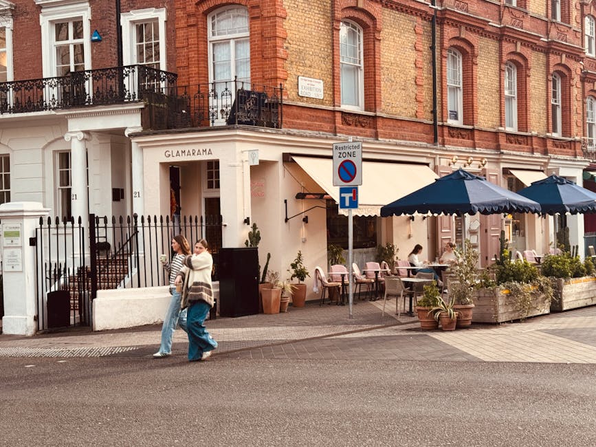 A street scene outside a building with a red brick facade and white architectural details, housing a café or restaurant with an outdoor seating area. The seating area is shaded by large navy blue umbrellas and decorated with potted plants, small trees, and a pink-painted wall. Three people are visible; two women are walking on the pavement near the entrance of the building, one of whom is wearing a striped top, and the other in a white sweater, while a third person sits at a table under the umbrellas, possibly enjoying a drink or meal. The sidewalk features a black metal fence, and a parking restriction sign is mounted on a pole in front of the establishment. The scene is well-lit with natural daylight, emphasizing the urban environment suitable for home relocation or furniture transport activities, which are often coordinated by Man with Van South Kensington during residential moving projects or removals in the area.