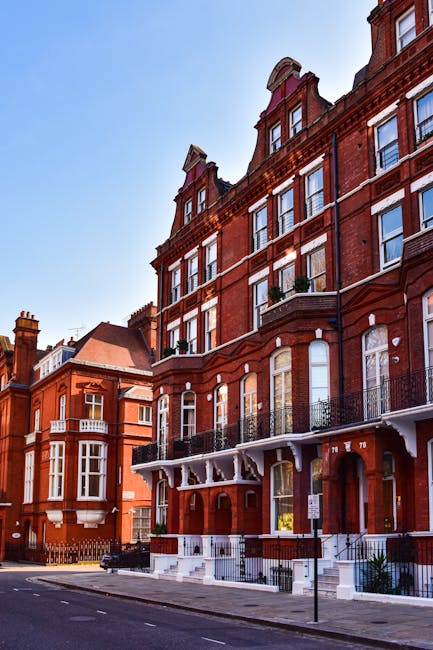 A row of historic red brick buildings in a residential area with large windows and decorative iron balconies on the upper floors. The ground floor features entry steps leading to doorways, some with small awnings and railings. The scene is taken during daylight under a clear blue sky, with a quiet street in front that has parked cars and a parked van used for home relocation services by Man with Van South Kensington. The van is partially visible on the left side, parked on the street pavement, and is likely being used for furniture transport or packing and moving activities related to house removals. The buildings and surroundings reflect a typical London neighbourhood, with clean streets and traditional architecture, supporting the context of house removals and relocation logistics.