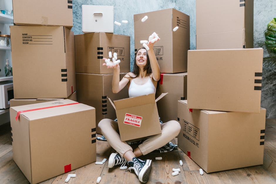 A young woman sitting cross-legged on a wooden floor inside a home, surrounded by numerous cardboard moving boxes of various sizes, some sealed with red tape and others open. She is holding and throwing white packing peanuts into the air while smiling slightly. The boxes are stacked against a textured grey wall with a small potted plant visible on the right side. The woman appears to be in the process of packing or unpacking during a house relocation, with some packing materials scattered on the floor. This scene depicts the packing and moving process associated with home removals services offered by Man with Van South Kensington, as indicated by the context of the page about moving costs in South Kensington.