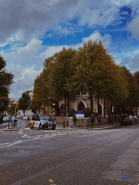 A residential street in South Kensington lined with parked cars on both sides, with several trees in full bloom displaying pink blossoms along the pavement. The street is flanked by elegant white terraced houses with detailed facades, large windows, and decorative elements. The scene is set during daytime with a partly cloudy sky overhead. This setting may illustrate a typical environment for home relocation or furniture transport, which Man with Van South Kensington's removals services could facilitate, highlighting the importance of careful vehicle parking, street access, and the scenic context of moving within this area.