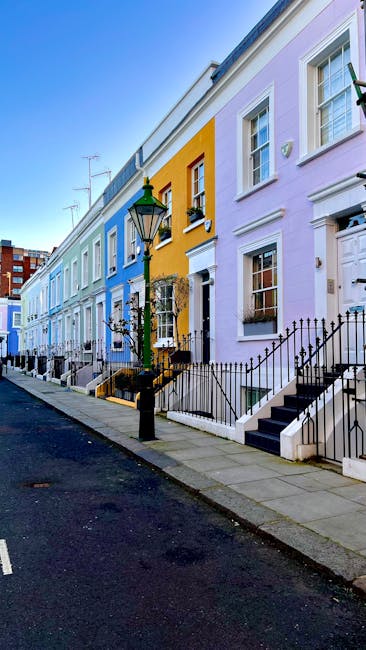 A row of colorful Victorian terraced houses in South Kensington, London, painted in pastel shades of blue, yellow, pink, and purple, with white window frames and decorative cornices. The pavement in front features black metal railings and low steps leading up to each front door. A green vintage-style street lamp is positioned on the sidewalk near a house with a black wrought iron gate. The sky above is clear and blue, with visible television antennas on some rooftops. This scene reflects an elegant residential area often associated with house removals and relocation services, as used by Man with Van South Kensington, with the environment implying a typical setting for furniture transport and home relocation activities.